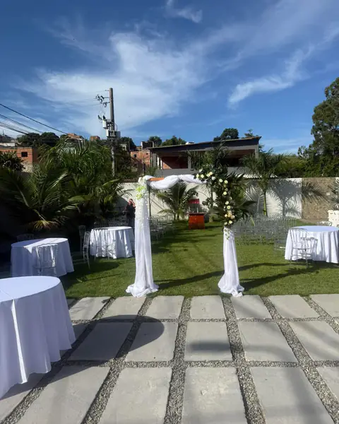 Arco de cerimônia com tecido branco e flores no gramado, entre mesas redondas com toalhas brancas.
